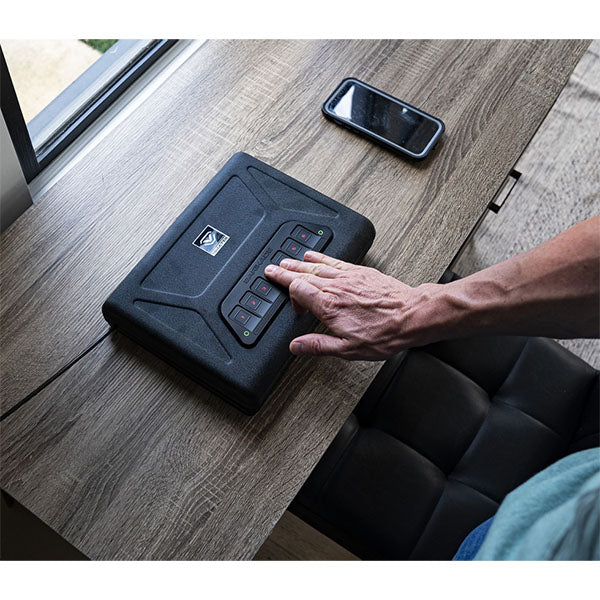 Hand tippt Code auf Tresor auf Holztisch, daneben liegt ein Smartphone.