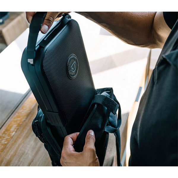 Person verstaut schwarzen Hartschalenkoffer in Rucksack, Innenbereich mit Holztisch.