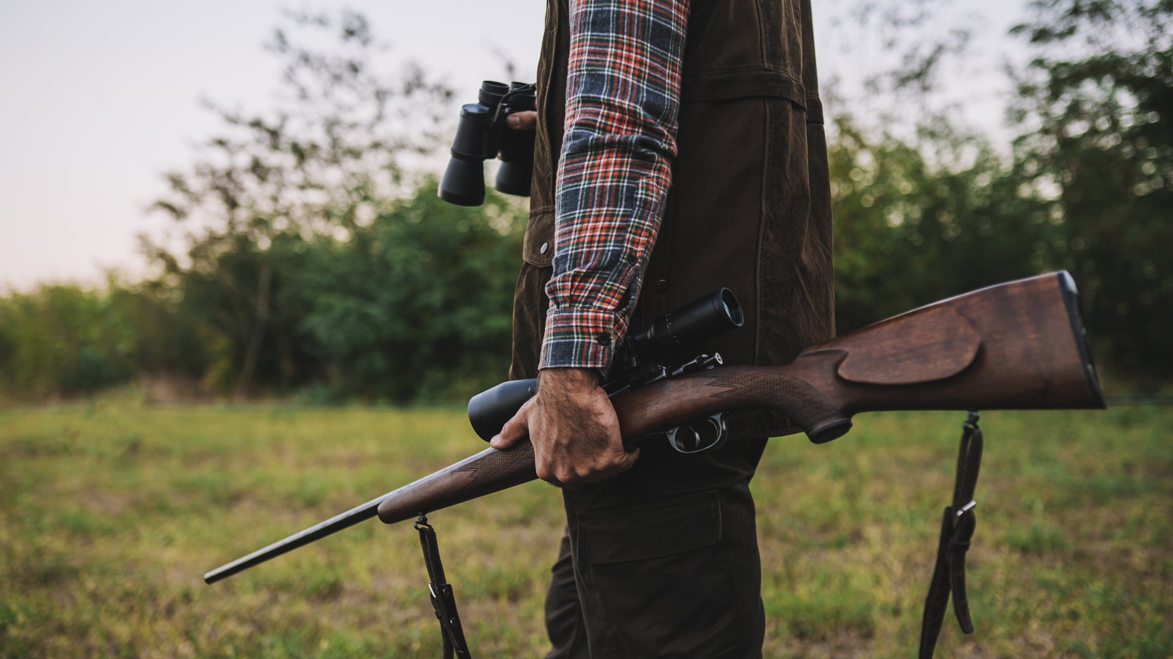 Jäger mit Gewehr und Fernglas steht auf einer Wiese am Waldrand.