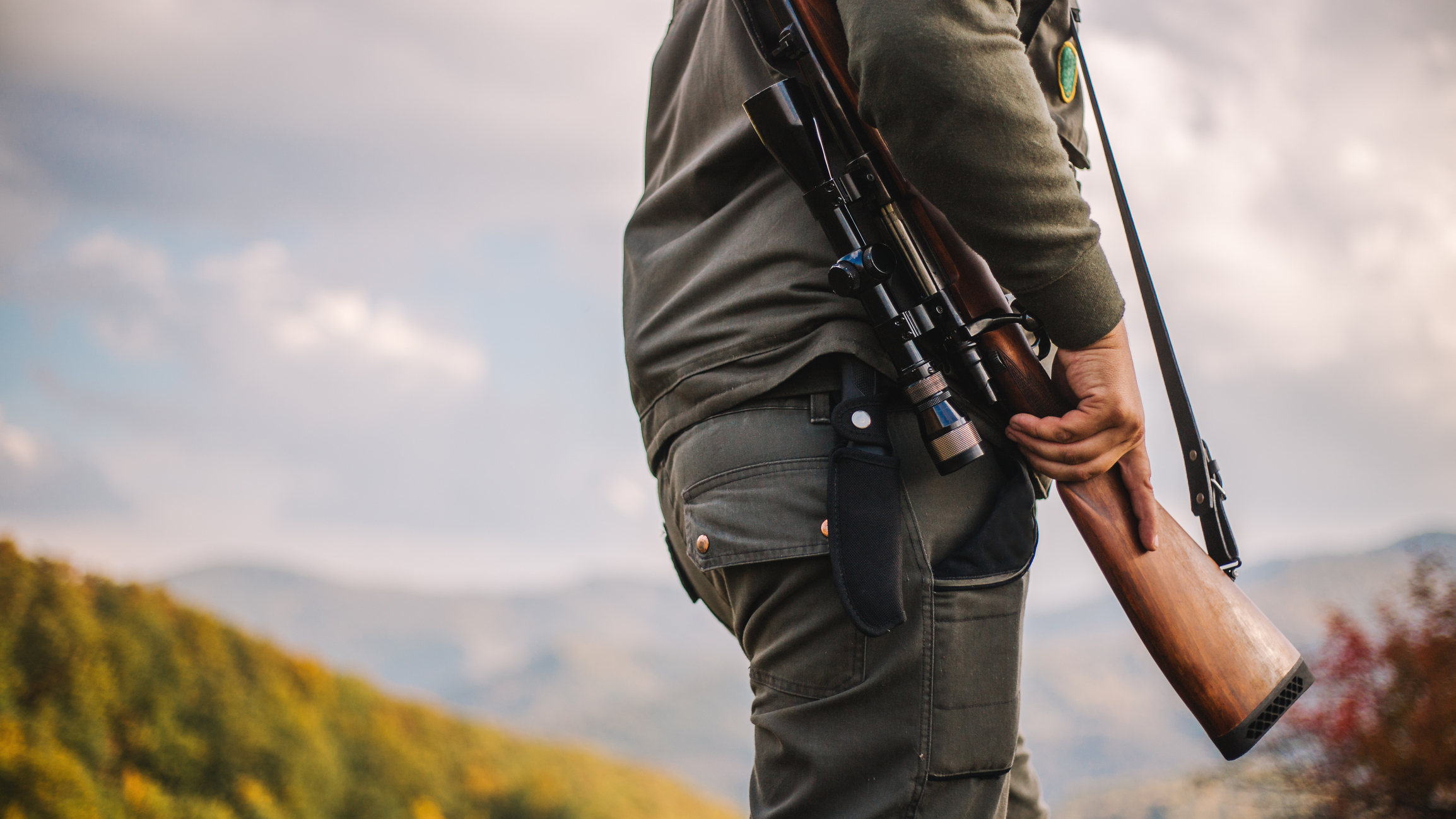 Person trägt Gewehr über der Schulter in bergiger Landschaft.