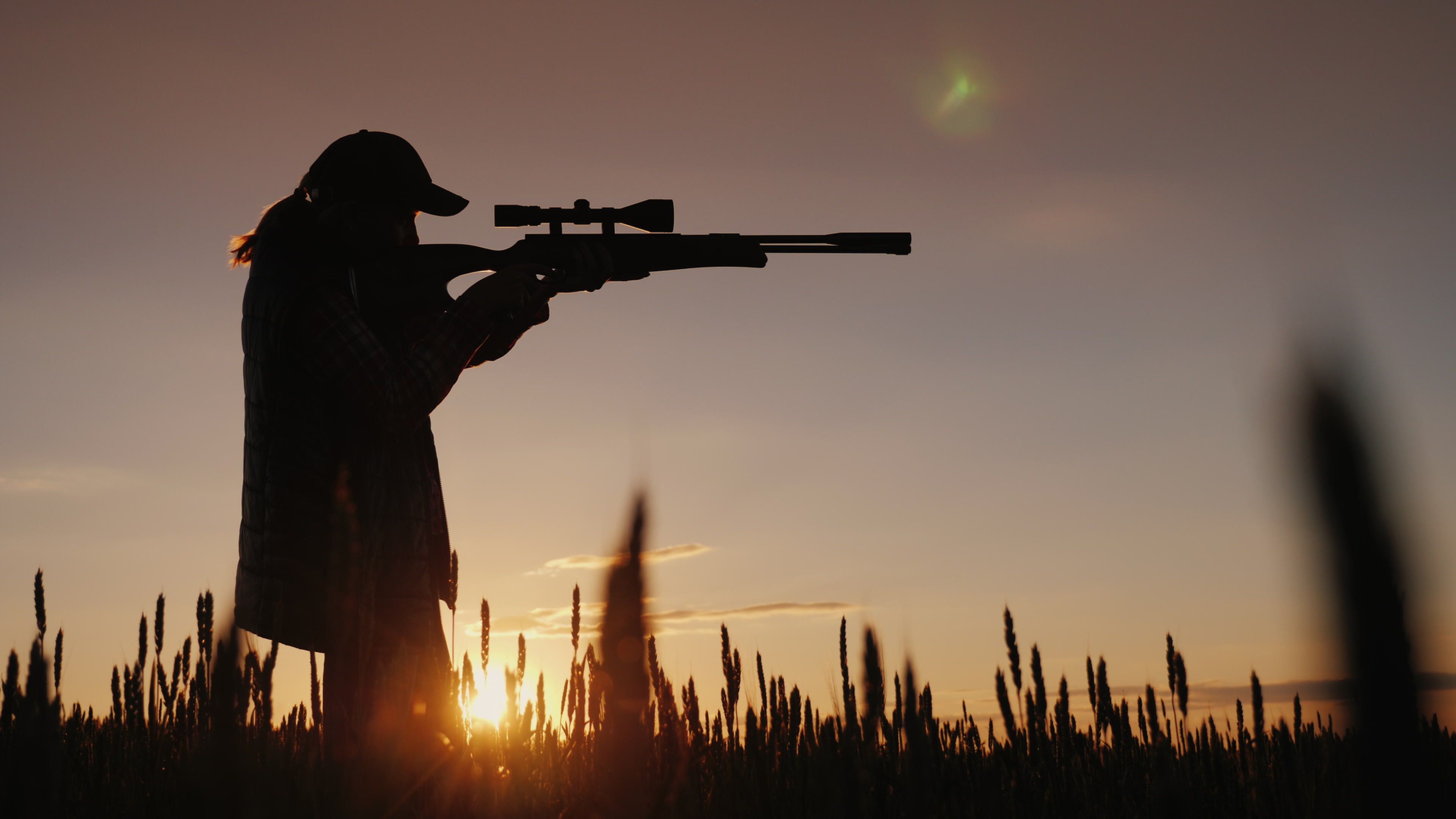 Person mit Gewehr im Feld bei Sonnenuntergang in seitlicher Silhouette.