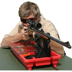 Person zielt mit Gewehr, liegend auf rotem Gestell, in Outdoor-Umgebung.